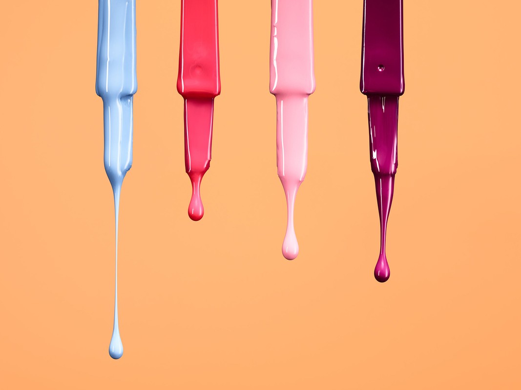 Four nail polish bottles with brushes dripping vibrant nail polish in blue, red, pink, and purple shades on a peach background.