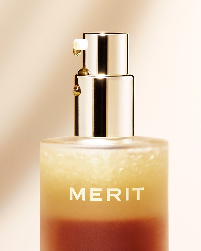 Close-up of a bottle of Merit beauty serum with a pump dispenser and golden liquid dripping from the nozzle, set against a beige background.