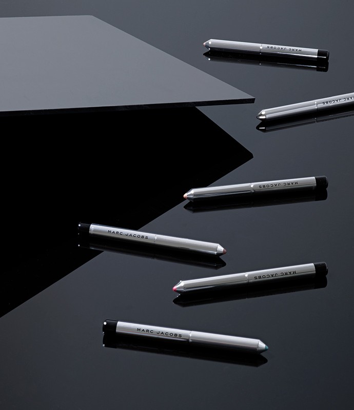 Several silver Marc Jacobs eyeliner pencils are displayed on a reflective black surface with a grey geometric shape in the background.