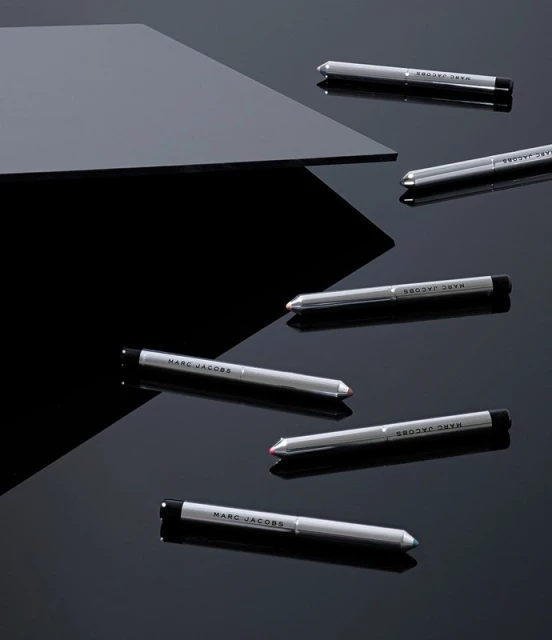 Several silver Marc Jacobs eyeliner pencils are displayed on a reflective black surface with a grey geometric shape in the background.