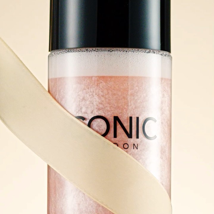Close-up of ICONIC LONDON setting spray bottle with black cap and cream-colored ribbon detail, against a soft, neutral background.