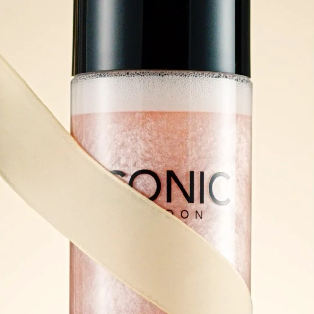 Close-up of ICONIC LONDON setting spray bottle with black cap and cream-colored ribbon detail, against a soft, neutral background.
