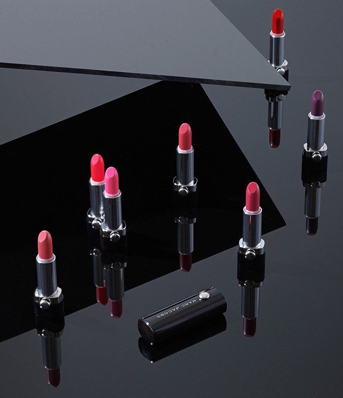 Product shot of several colorful lipsticks in silver and black tubes, arranged on a glossy black surface with geometric reflections.