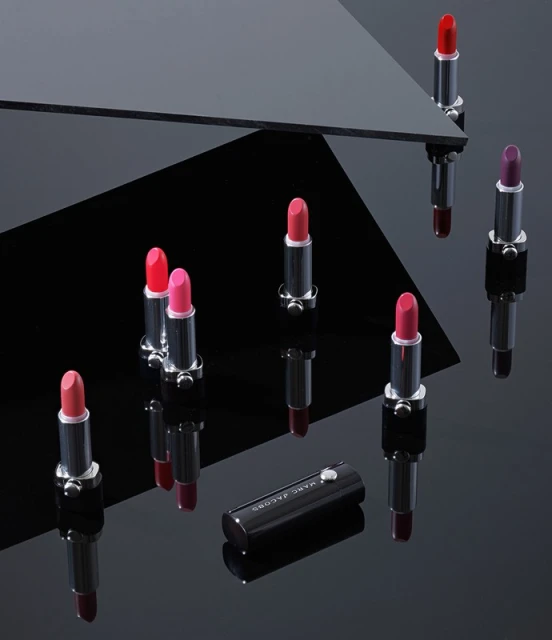 Product shot of several colorful lipsticks in silver and black tubes, arranged on a glossy black surface with geometric reflections.