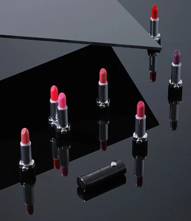 Product shot of several colorful lipsticks in silver and black tubes, arranged on a glossy black surface with geometric reflections.