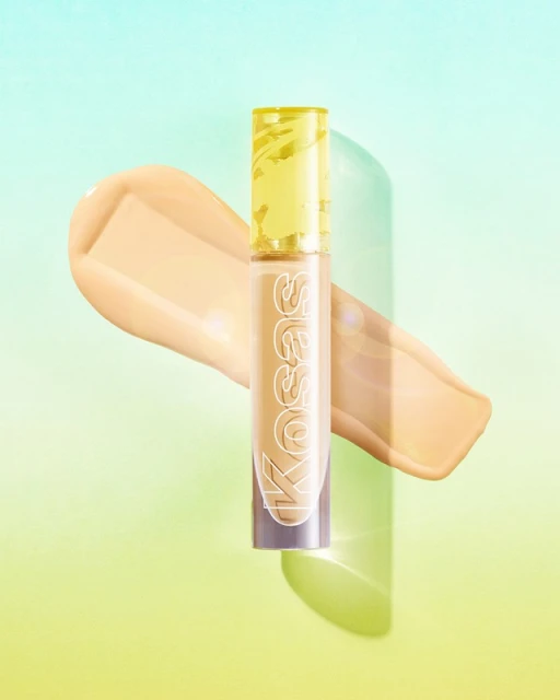 Kosas Revealer Concealer tube with yellow lid and beige concealer swatch on a green and blue gradient background in a studio shot.