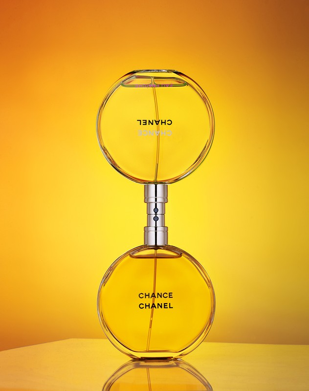 Close-up studio shot of two Chanel Chance perfume bottles stacked, showcasing the golden liquid and circular glass design against a warm gradient background.