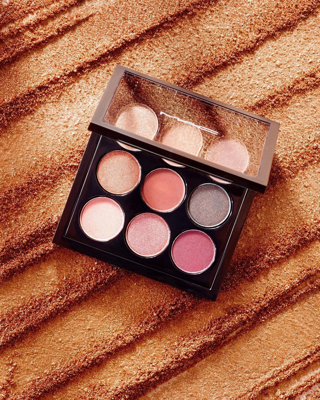Overhead studio shot of an open eyeshadow palette with multiple shades of pink, brown, and grey, set against a textured golden background.