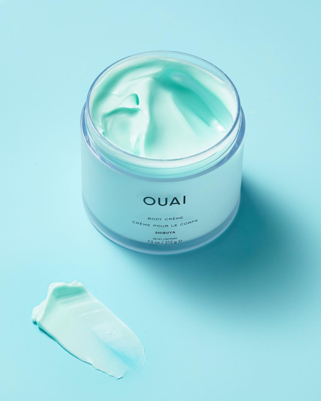 Elevated view of OUAI body cream in a clear jar and a smear of cream on a blue background, showcasing beauty and skincare.