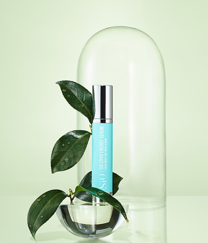 Still life of SiO Cryo Energy Serum bottle with green leaves, water drops, and clear glass dome on a light green background.