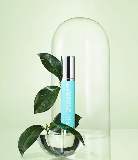 Still life of SiO Cryo Energy Serum bottle with green leaves, water drops, and clear glass dome on a light green background.