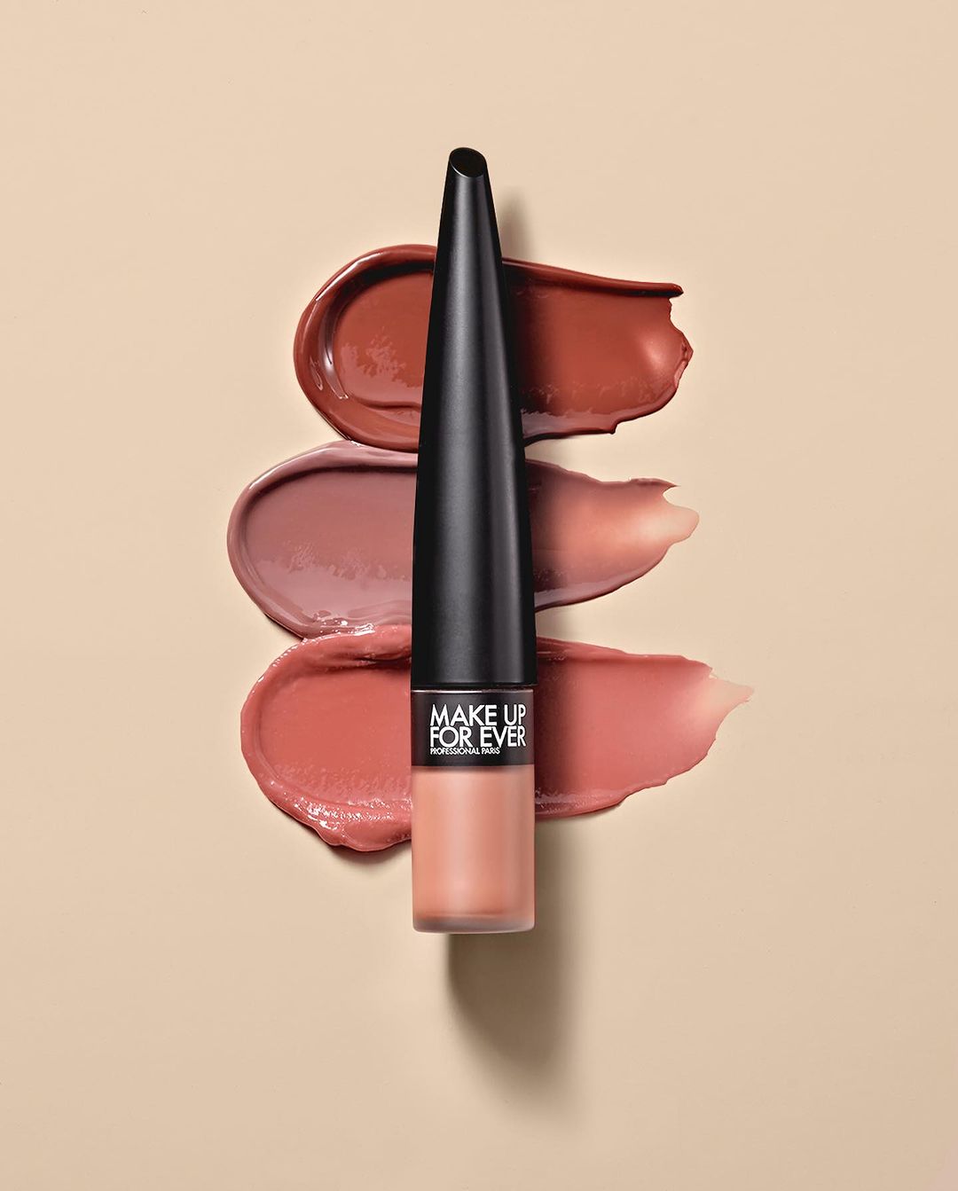 Lip product tube standing vertically centered over three horizontal cosmetic swatches on beige background