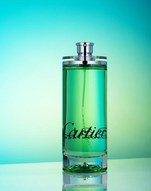 Cylindrical transparent glass bottle of green liquid perfume with silver top and black 'Cartier' logo against a teal and light-green backdrop.