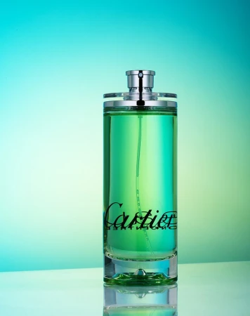 Cylindrical transparent glass bottle of green liquid perfume with silver top and black 'Cartier' logo against a teal and light-green backdrop.