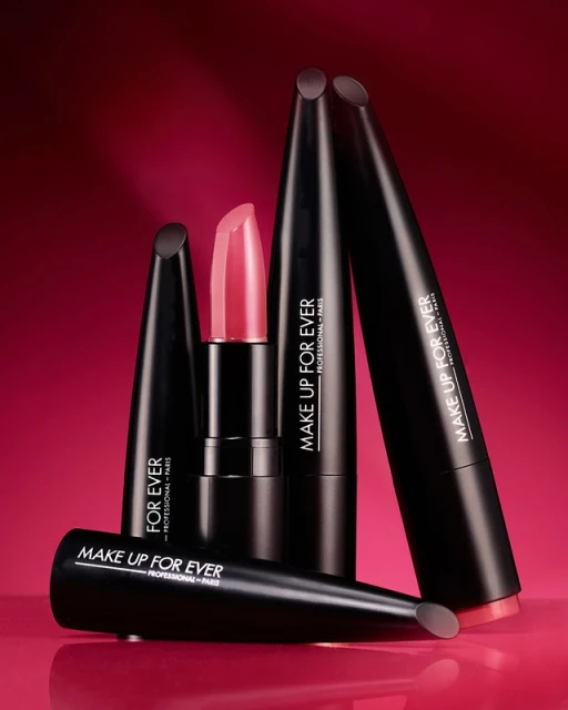 Close-up studio shot of Make Up For Ever lipsticks in sleek black packaging with a pink background, showcasing beauty and cosmetic products.