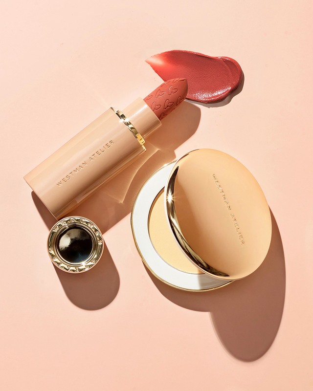 Close-up shot of Westman Atelier makeup products including lipstick, compact, and dab of lipstick color on a pale pink background.
