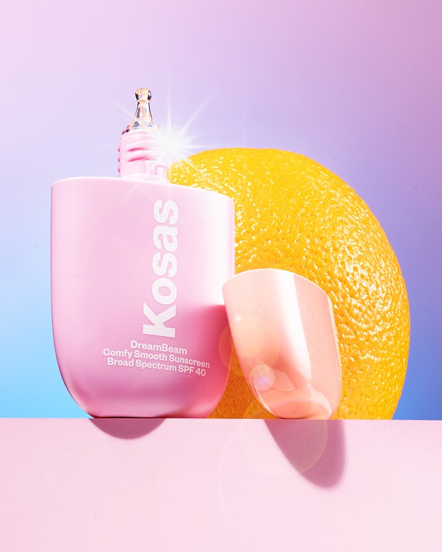 Product shot of a bottle of Kosas DreamBeam sunscreen with an orange and a translucent stand on a pink surface against a gradient background.