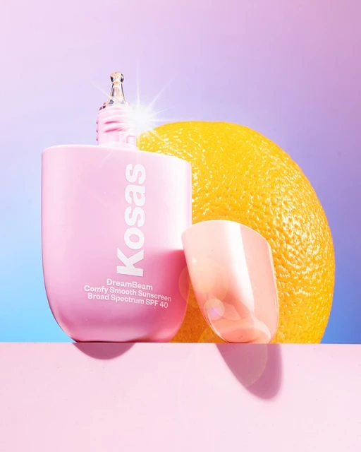 Product shot of a bottle of Kosas DreamBeam sunscreen with an orange and a translucent stand on a pink surface against a gradient background.