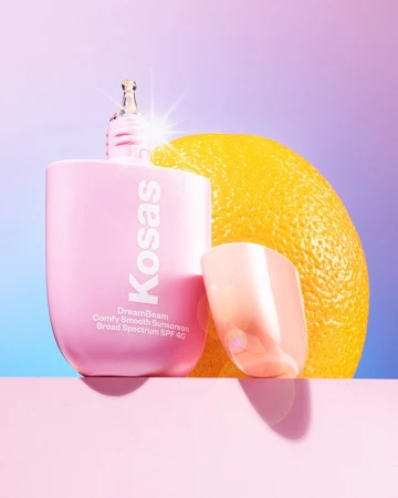 Product shot of a bottle of Kosas DreamBeam sunscreen with an orange and a translucent stand on a pink surface against a gradient background.