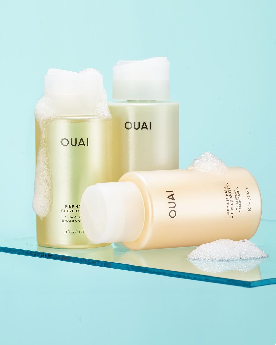 Three bottles of Ouai shampoo on a clear surface against a blue backdrop, two with white foam on top, in a beauty product shot.