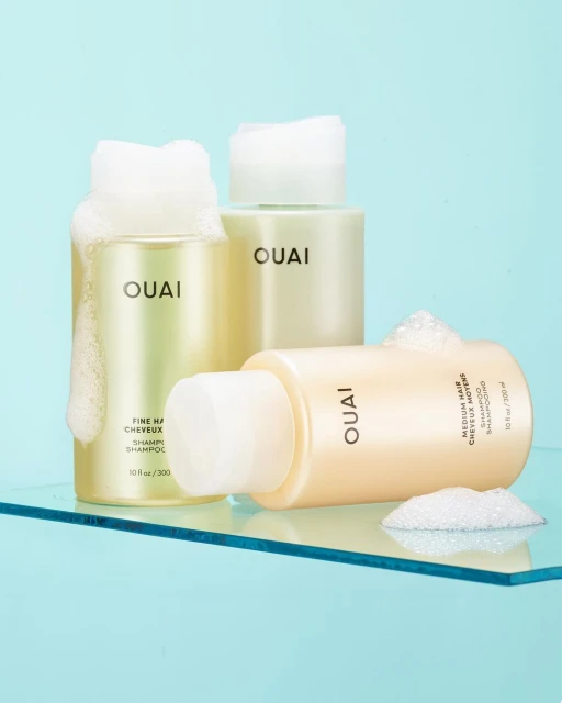 Three bottles of Ouai shampoo on a clear surface against a blue backdrop, two with white foam on top, in a beauty product shot.