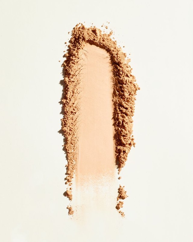 Tan cosmetic powder swatch on a white surface, illuminated by bright, diffused lighting. The powder has a fine, granular texture.