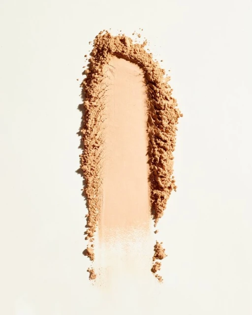 Tan cosmetic powder swatch on a white surface, illuminated by bright, diffused lighting. The powder has a fine, granular texture.
