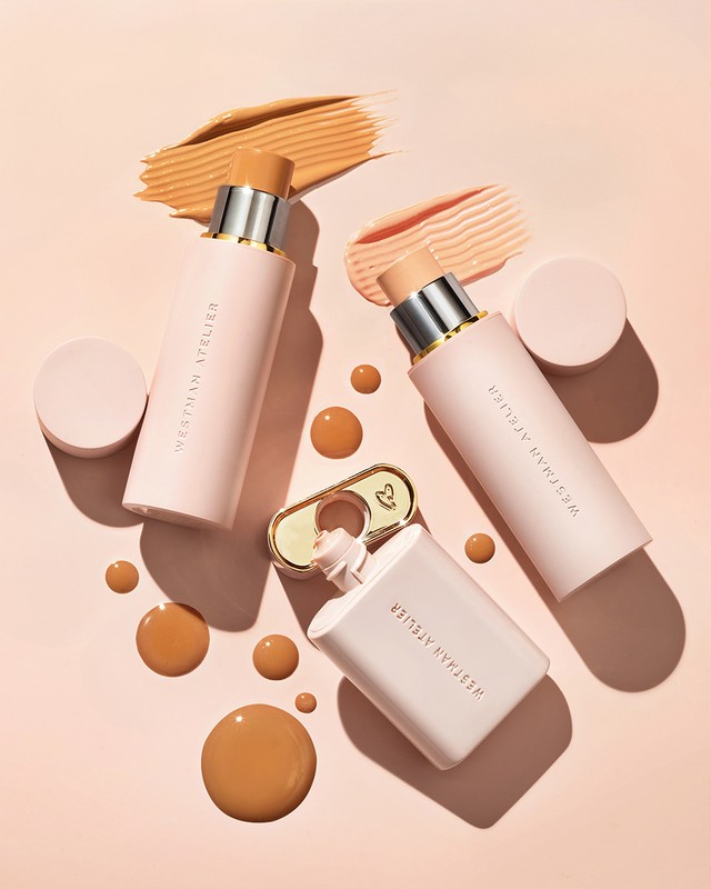Several pink-toned Westman Atelier makeup products are arranged on a light pink surface with makeup splotches and drops.