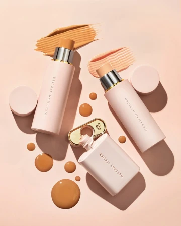 Several pink-toned Westman Atelier makeup products are arranged on a light pink surface with makeup splotches and drops.