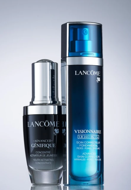 Two Lancome skincare bottles, Génifique and Visionnaire, stand in a product shot with blue, black and silver design elements on a light gradient background.