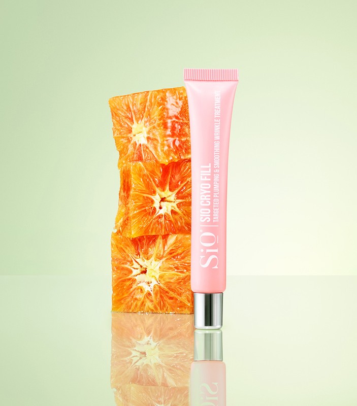 Studio shot of a pink lotion tube next to a stacked pile of sliced oranges against a gradient green background. Focus on skincare and beauty.