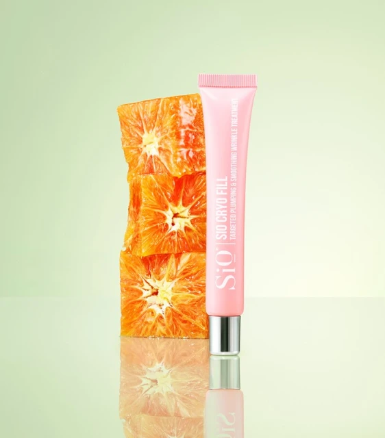 Studio shot of a pink lotion tube next to a stacked pile of sliced oranges against a gradient green background. Focus on skincare and beauty.