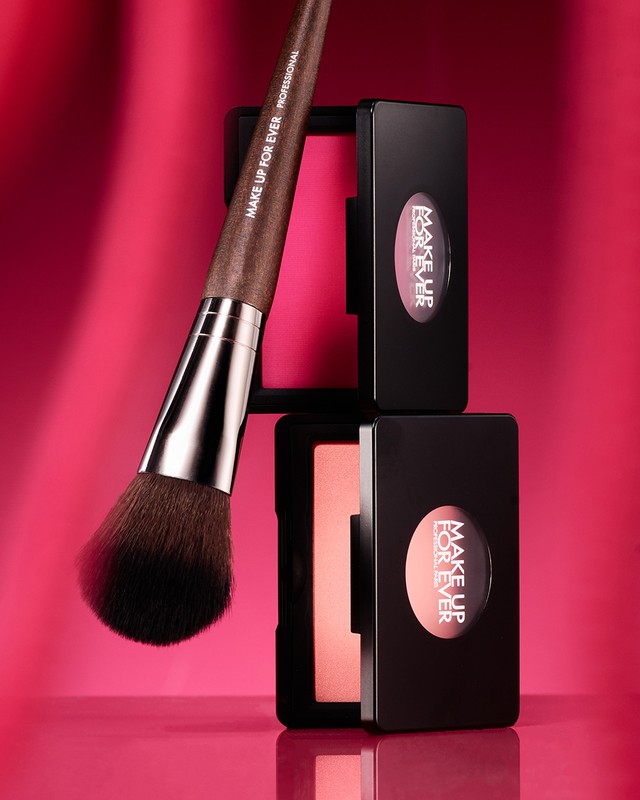 Blush brush alongside Make Up For Ever blush compacts on a reflective surface with a pink background and studio lighting.