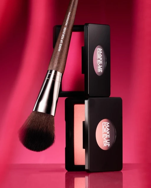 Blush brush alongside Make Up For Ever blush compacts on a reflective surface with a pink background and studio lighting.