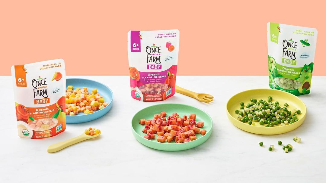 Several pouches and bowls of Once Upon a Farm baby food displayed on a white table against a peach-colored background. Healthy meal options.