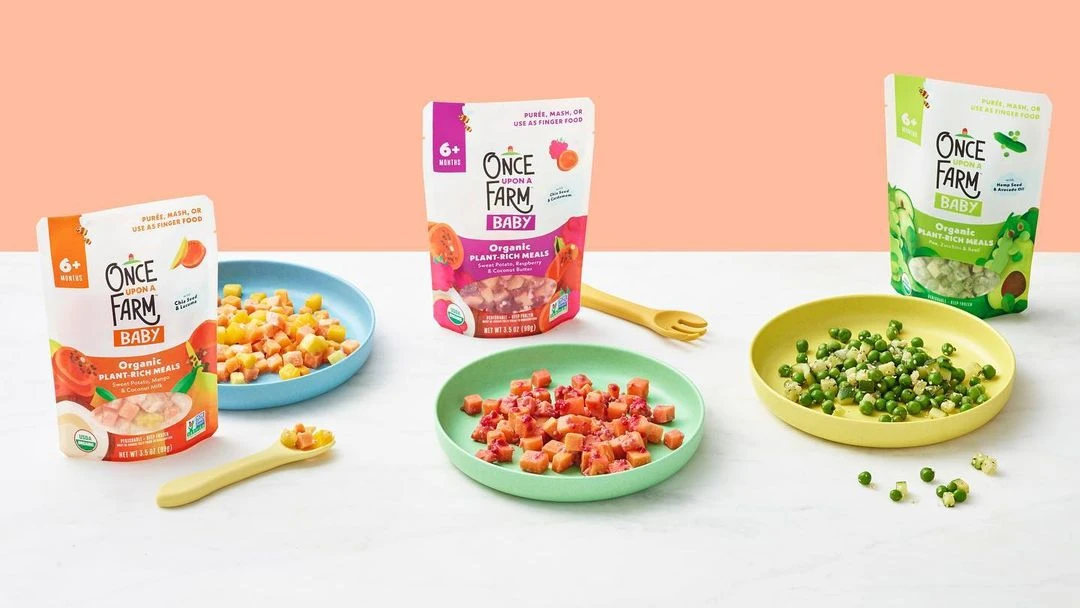 Several pouches and bowls of Once Upon a Farm baby food displayed on a white table against a peach-colored background. Healthy meal options.