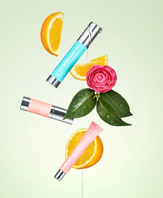 Cosmetic products from skincare and beauty brands floating with orange slices and pink flower on light green background in studio shot.