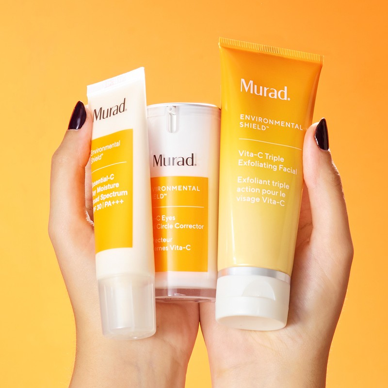 Close-up of fair-skinned hands holding Murad skincare products in white and orange tubes and pump bottles on a yellow-orange backdrop.