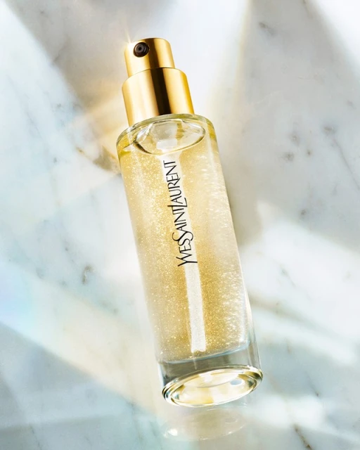 Close-up product shot of Yves Saint Laurent shimmer body oil in a clear glass bottle with a gold cap on a marble surface.
