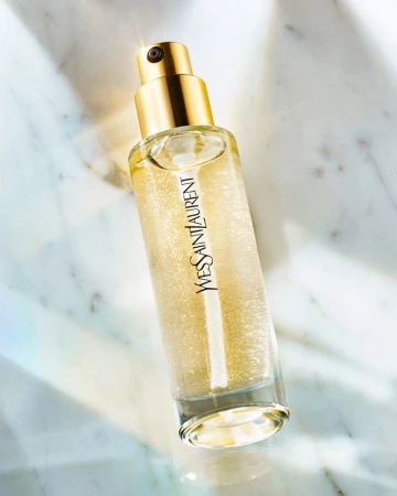 Close-up product shot of Yves Saint Laurent shimmer body oil in a clear glass bottle with a gold cap on a marble surface.