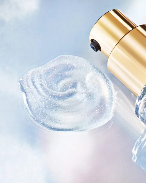 Close-up of a clear skincare serum swatch with shimmering flecks, next to a gold pump bottle against a soft blue background.