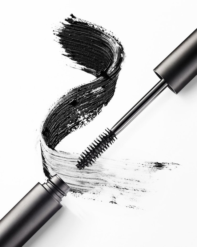 Close-up of a mascara wand with black mascara on the bristles next to a swirl of mascara on a white surface.
