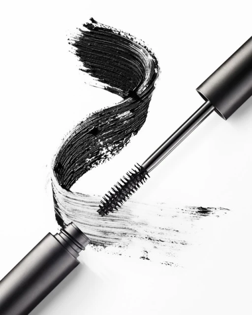 Close-up of a mascara wand with black mascara on the bristles next to a swirl of mascara on a white surface.