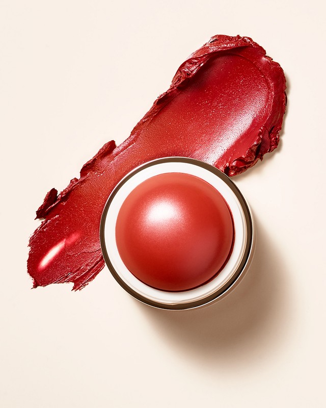Close-up of a red-hued cream makeup stick with a swatch on a beige surface, showcasing the product's color and creamy texture.