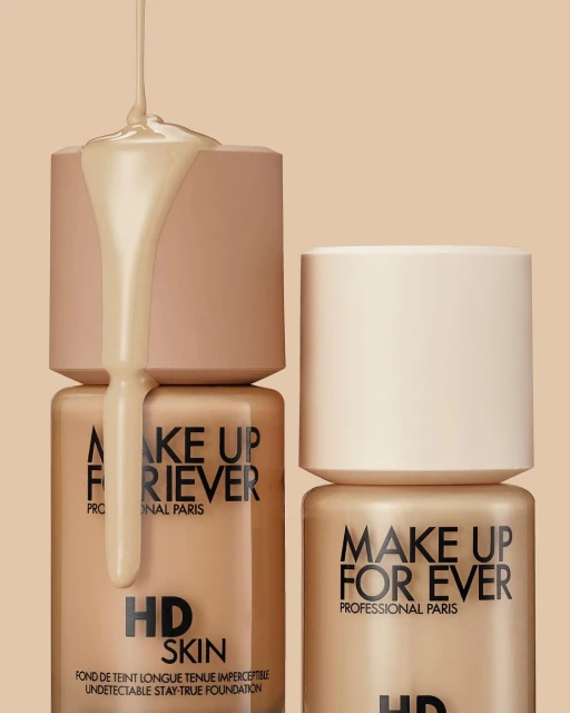 Cosmetic still life: Makeup Forever HD Skin foundation bottles are displayed with foundation liquid dripping over one of the bottles.