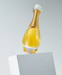 Amphora-shaped perfume bottle with gold neck and spherical cap resting diagonally on a grey block against a white gradient background