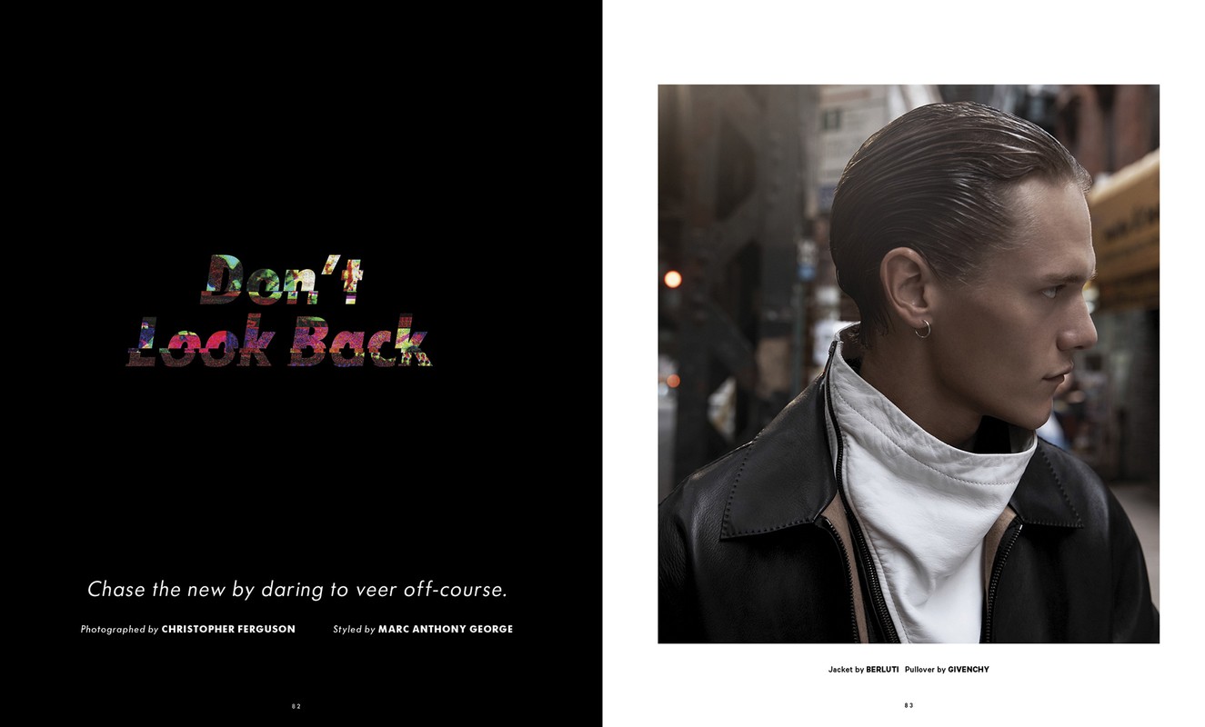 Fashion editorial shot of adult male model with slicked-back hair, wearing a black leather jacket and a white pullover against an urban background.