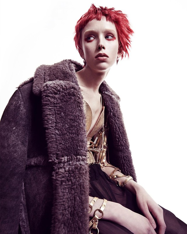 A woman with short red hair poses in a photo studio wearing a metallic gold top, dark shearling coat, and gold chain jewelry.