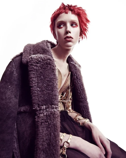 A woman with short red hair poses in a photo studio wearing a metallic gold top, dark shearling coat, and gold chain jewelry.
