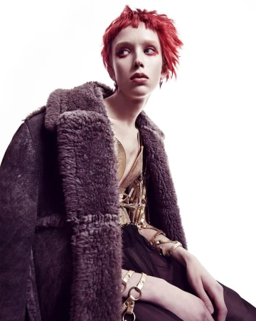 A woman with short red hair poses in a photo studio wearing a metallic gold top, dark shearling coat, and gold chain jewelry.